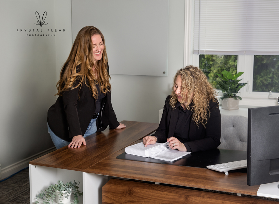 Two women smiling and collaborating in a modern office space—one seated at a desk reviewing notes, the other standing beside her in conversation. Natural light, indoor branding photography.